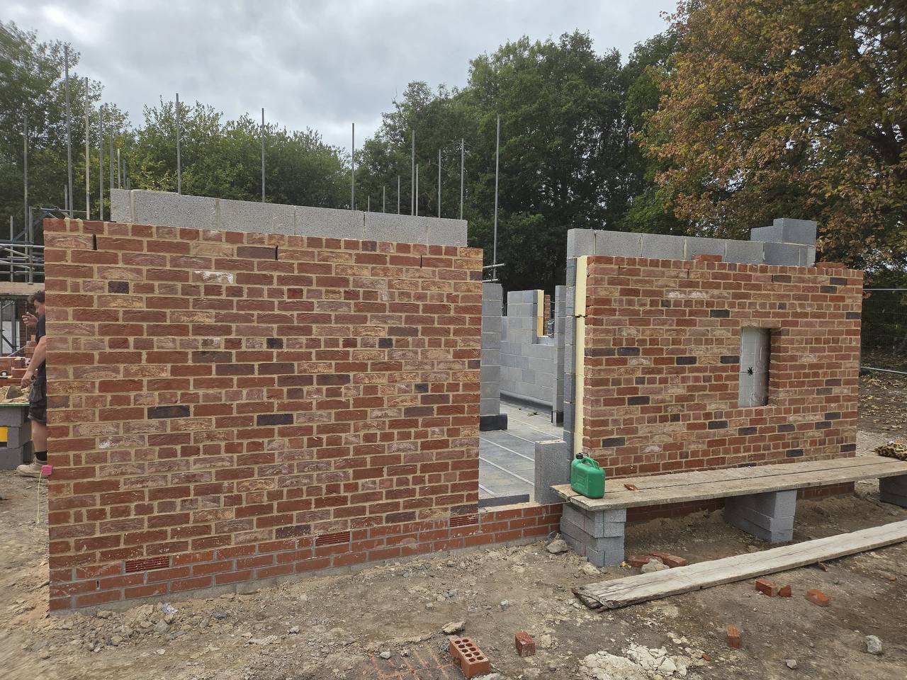 Two new-build brick cavity walls under construction with blockwork cores and wall ties, set on a working site with scaffolding and trees behind
