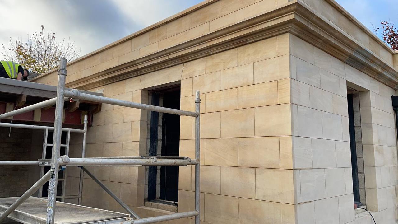New build ashlar sandstone pavilion nearing completion, with a moulded stone cornice, precisely jointed coursing and dressed openings, viewed past the working scaffold